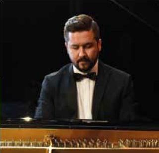 Pianista Jacek Obstarczyk gra na fortepianie, ubrany jest w czarny smoking, białą koszulę oraz czarną muszkę
