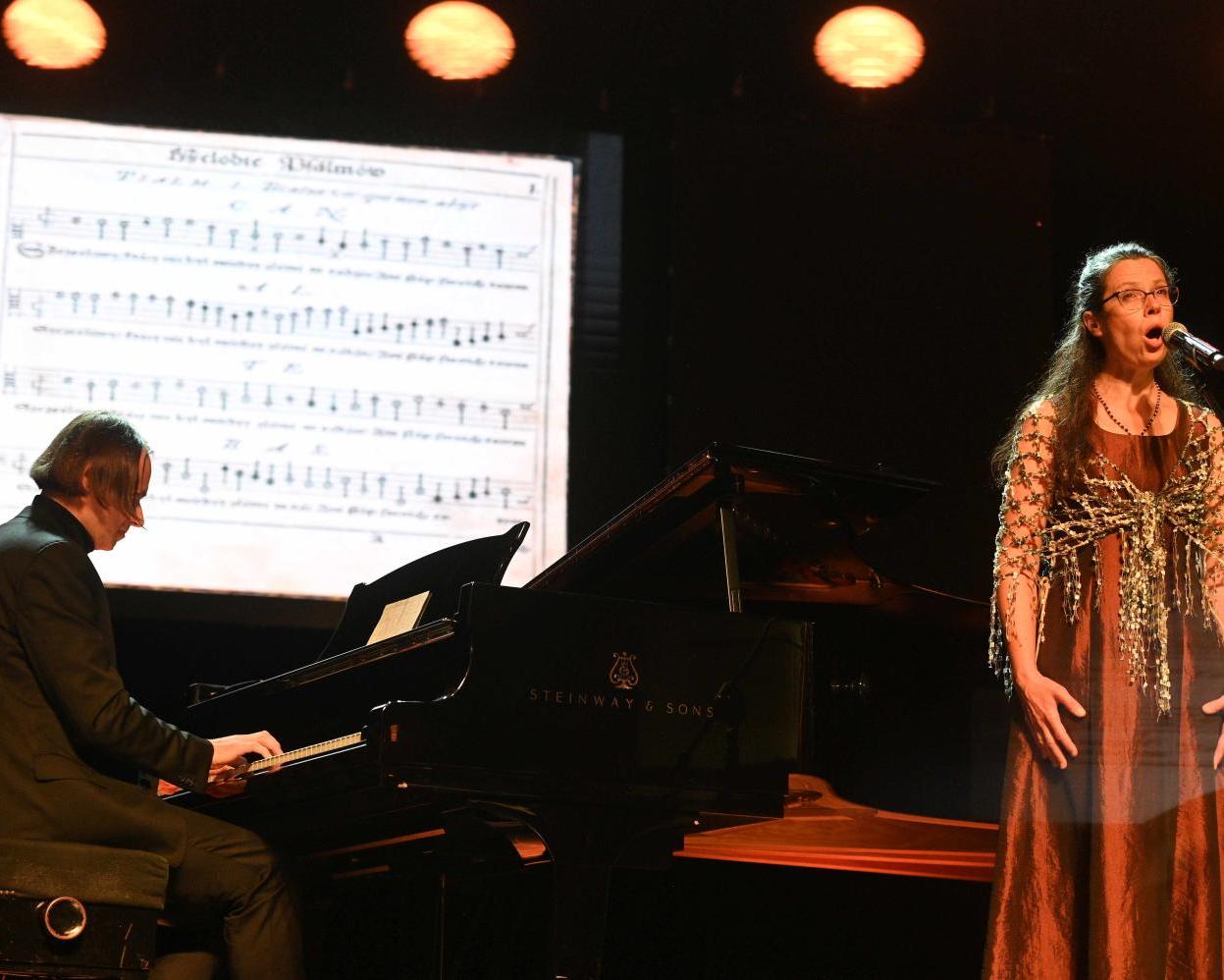 Śpiewaczka Joanna Korpiela-Jatkowska wykonuje utwór z organistą Tomaszem Orlowem grającym na czarnym fortepianie, w tle nuty starego utworu