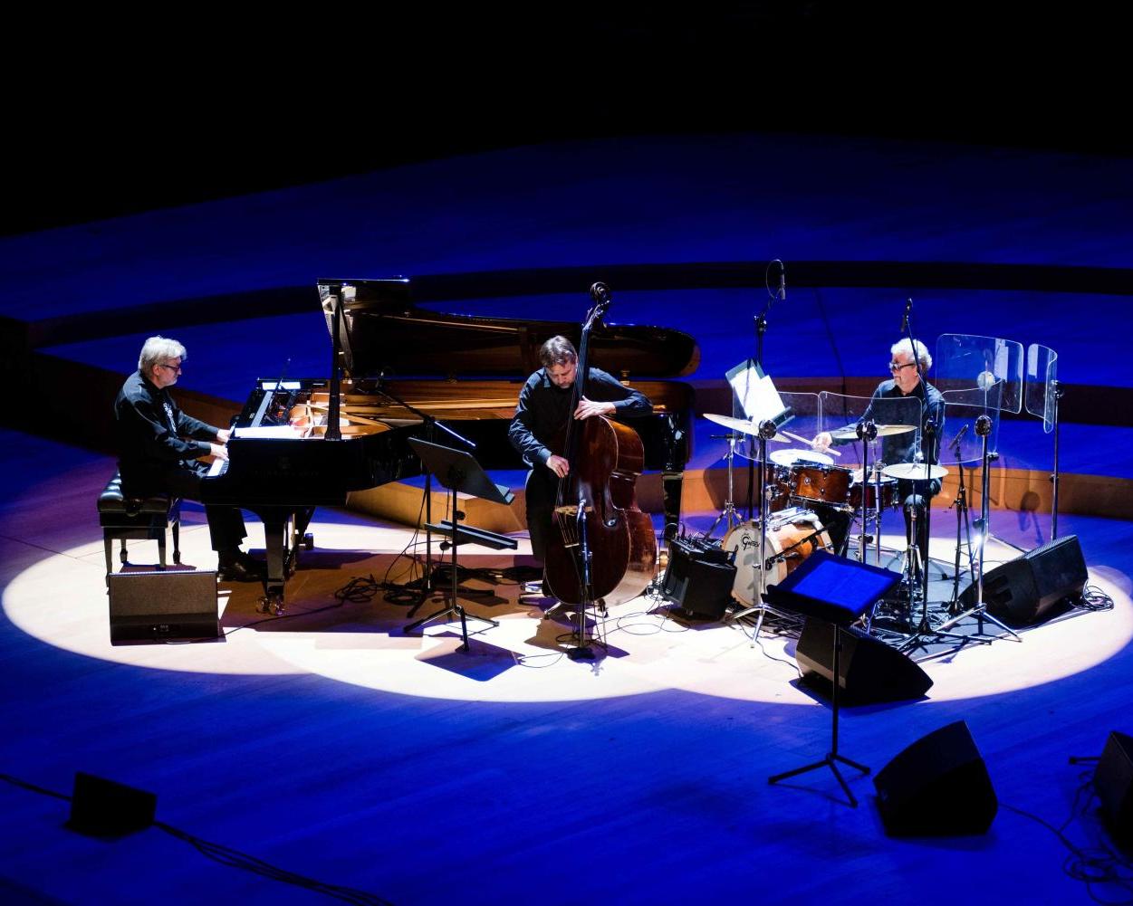 Trio Andrzeja Jagodzińskiego, pianista siedzi przy czarnym fortepianie, kontrabasista stoi z instrumentem, perkusista siedzi za składem instrumentów