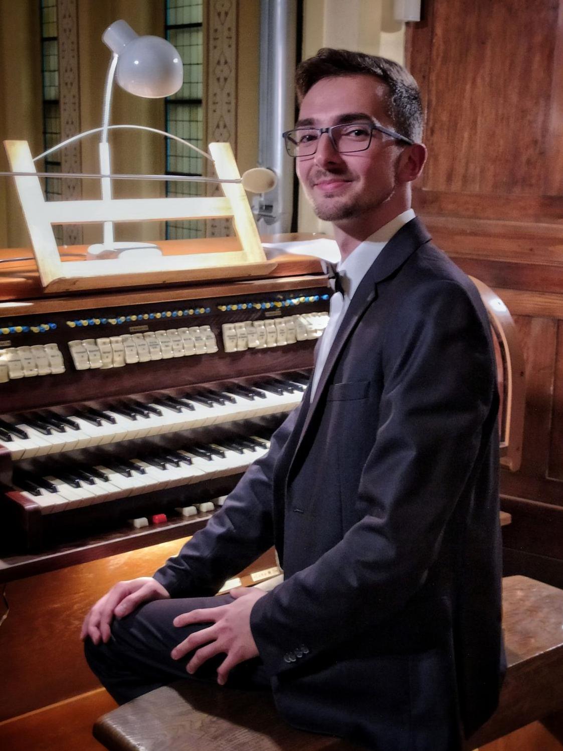 Organista Maciej Leśniak siedzi przy kontuarze instrumentu, ubrany jest w garnitur i muszkę