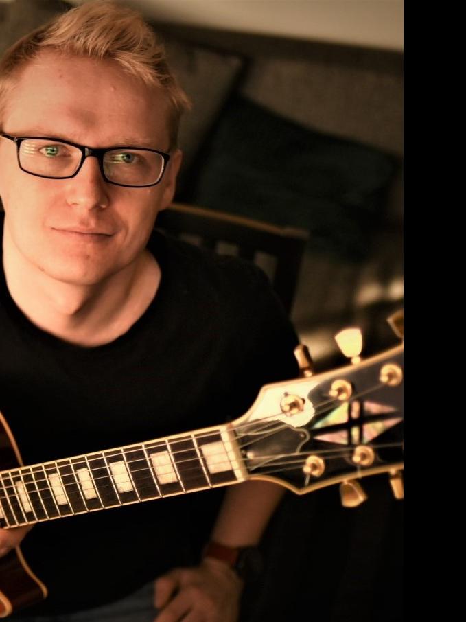 Gitarzysta Przemysław Hanaj gra na instrumencie, widoczne są struny i pudło rezonansowe gitary w kolorze brązowym, artysta nosi okulary i ubrany jest w czarną koszulkę